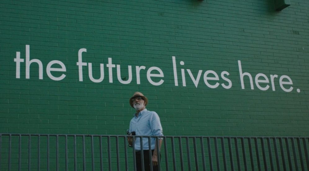 A bearded man in front of a green brick wall that reads "the future lives here."
