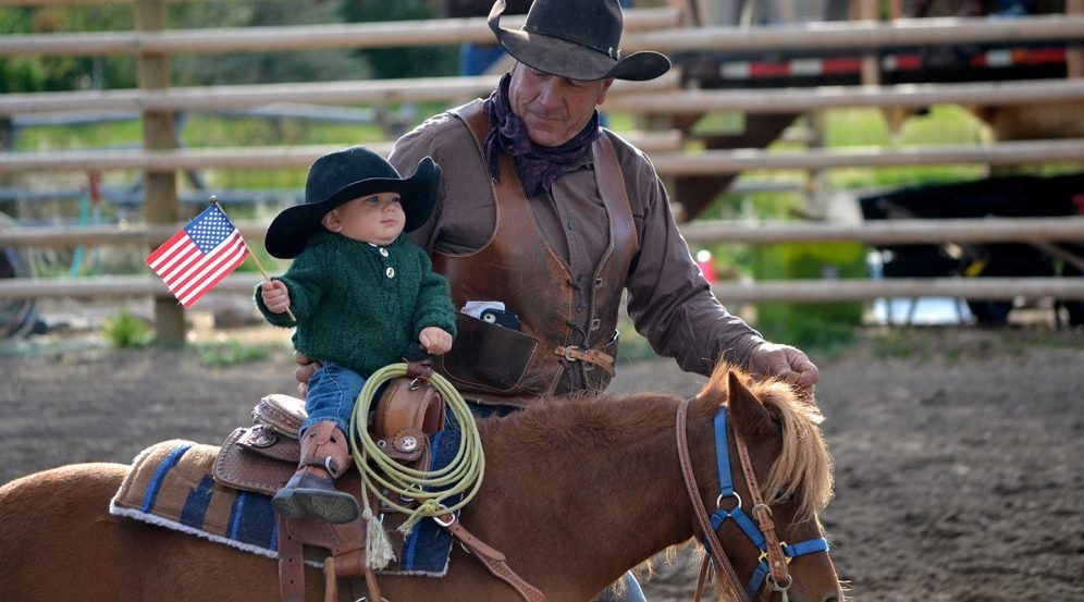 A baby in a cowboy hat riding a horse with a member of staff at The Ranch At Rock Creek. Shorter image.